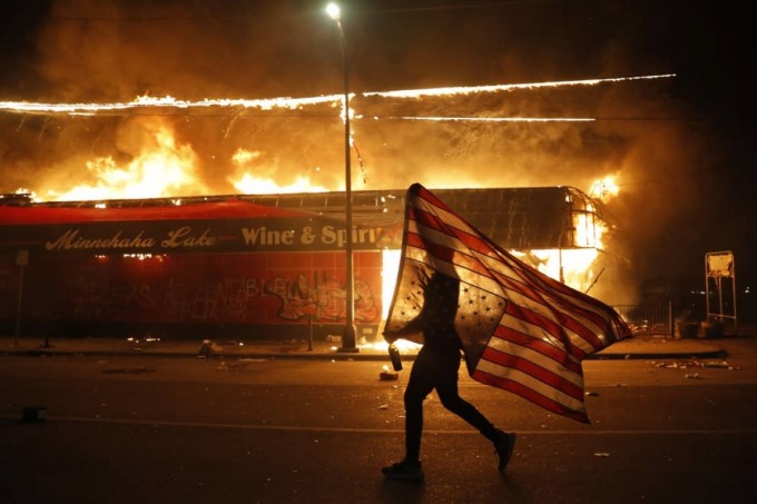 Một người biểu tình cầm ngược lá cờ Mỹ thể hiện sự đau khổ, bên cạnh một tòa nhà đang bốc cháy vào ngày 28/5 ở Minneapolis, sau khi cảnh sát ghì chết George Floyd. Ảnh: AP
